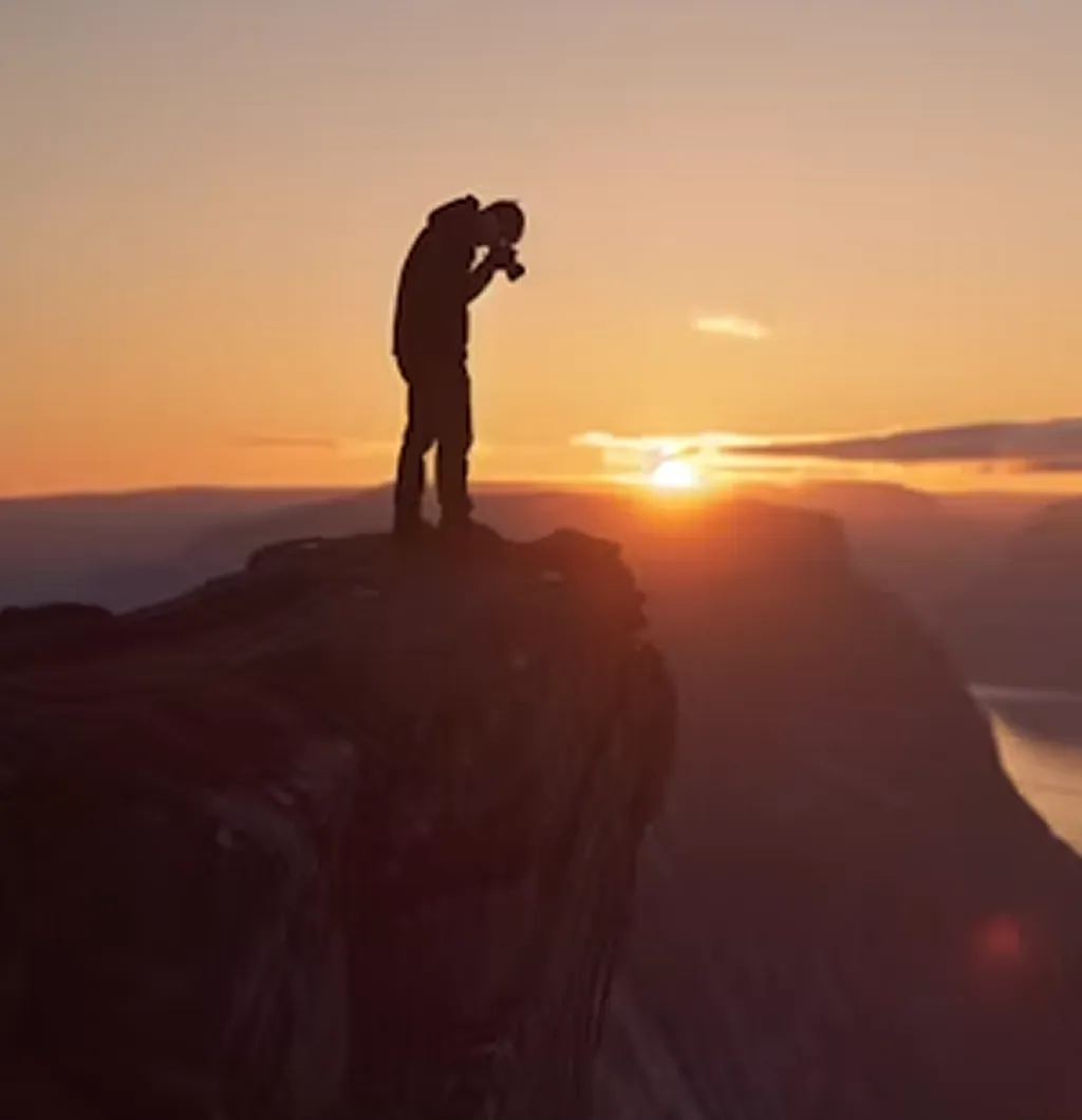 Silhouette of a person taking a photo on a cliff during a sunset with mountains in the background.