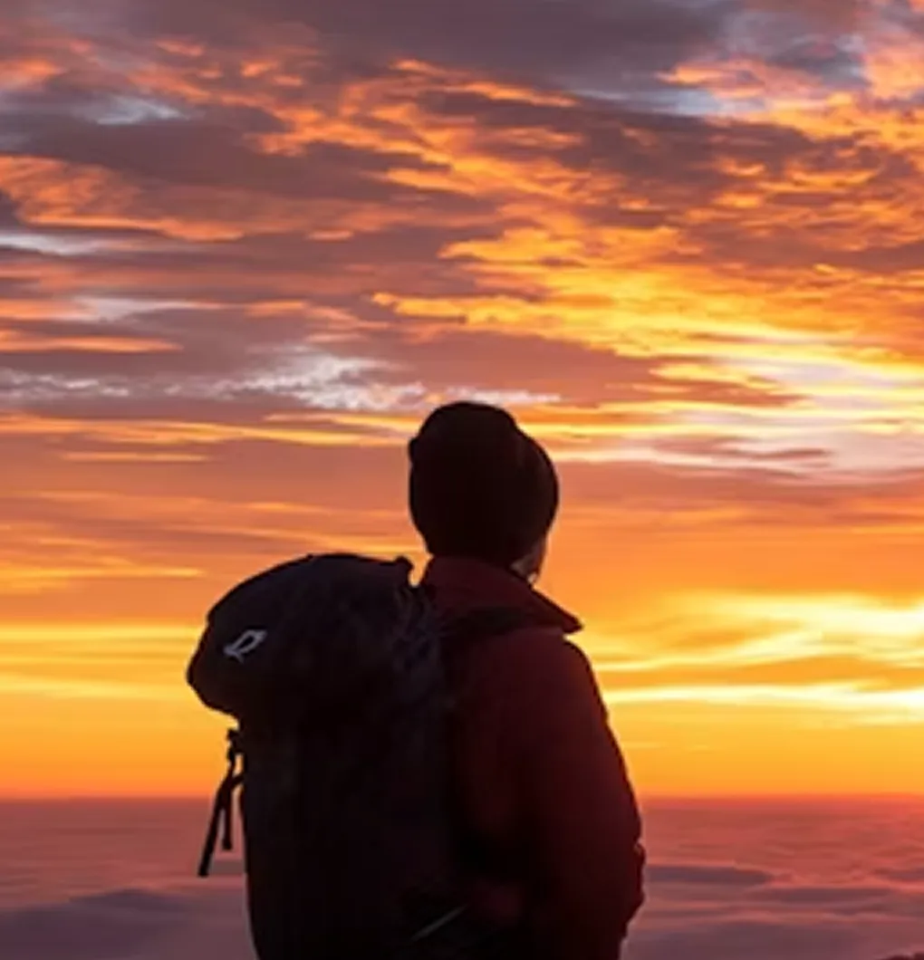 Person with backpack watches colorful orange and yellow sunset above clouds.