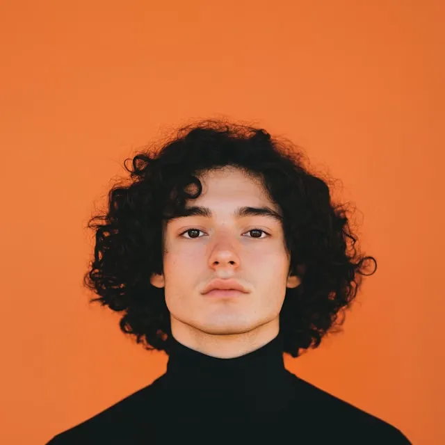 Young person with curly black hair wearing a black turtleneck against an orange background.
