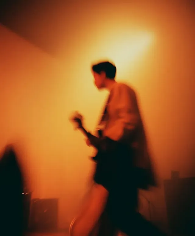 Blurred silhouette of a person playing electric guitar against warm orange background.