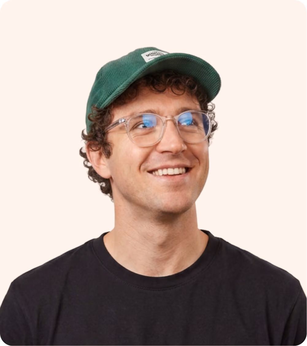 Smiling partner marketing manager with curly hair, clear glasses, green cap, and black shirt on light beige background.