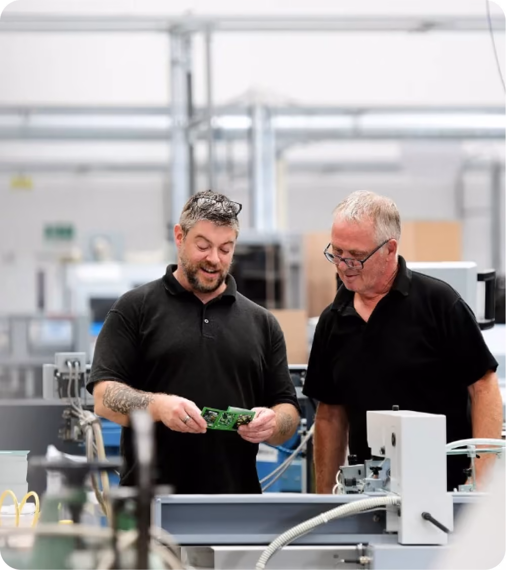 Two factory workers inspecting a green circuit board in an electronics manufacturing setting.