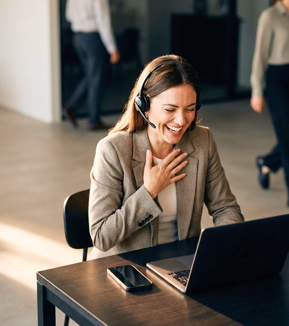 Woman wearing headset laughs while video calling on a laptop at a desk with a smartphone.