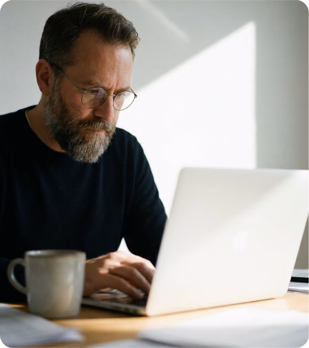 Bearded man wearing glasses working on a laptop at a desk with a mug nearby.