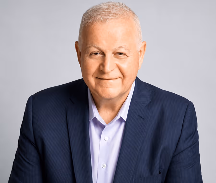 Portrait of a smiling older man in a dark blazer and light blue shirt against a plain background.