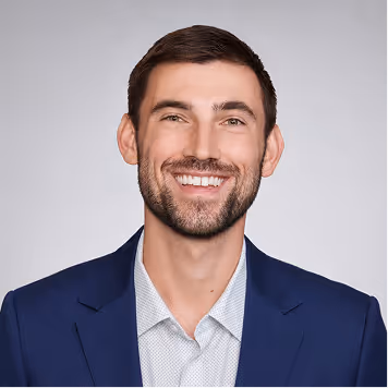 Smiling man with short dark hair and beard wearing a navy suit jacket and white shirt.
