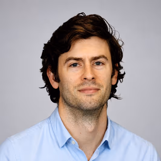 Headshot of a man with dark wavy hair wearing a light blue collared shirt against a gray background.