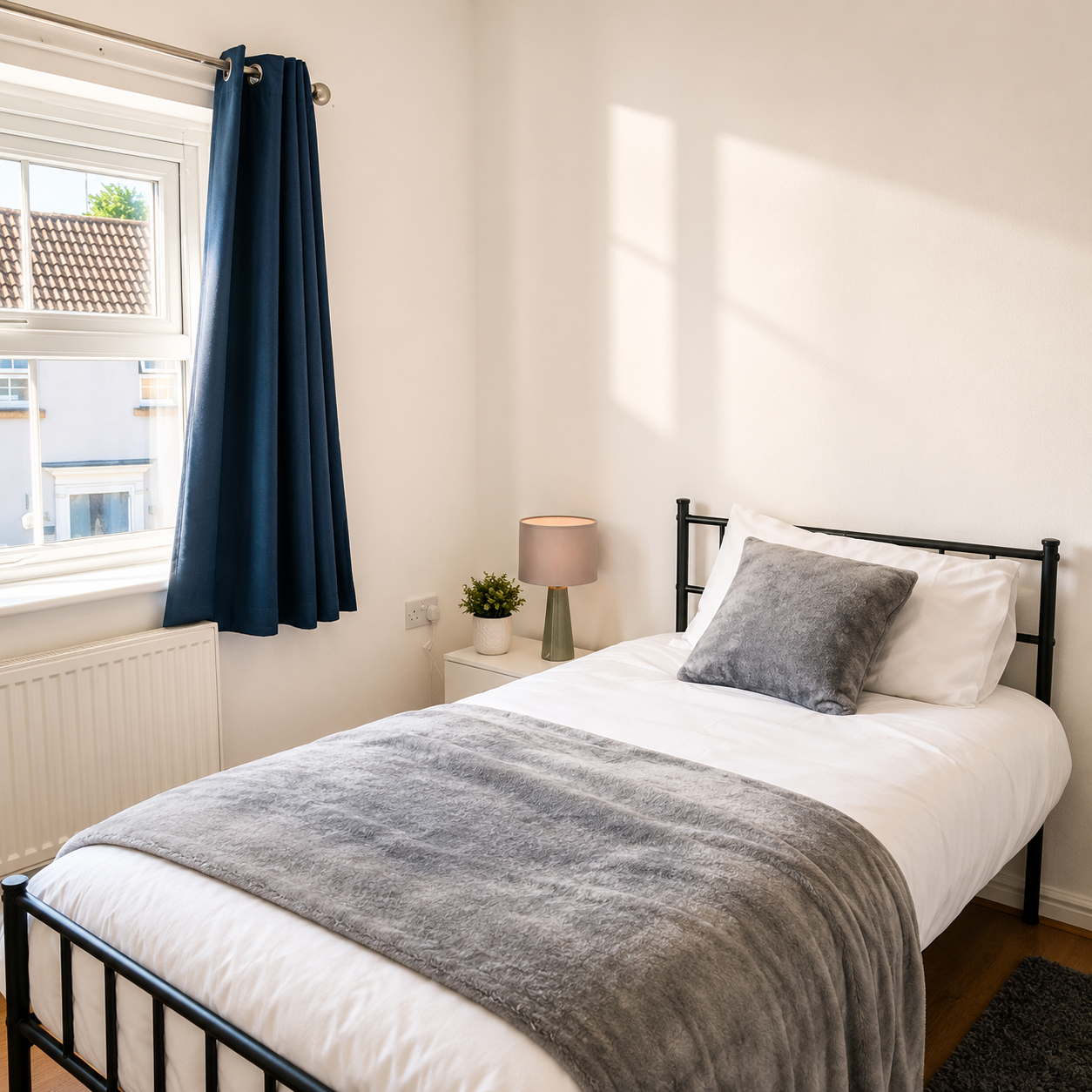 Single bed with gray blanket and pillow in a room with navy curtain and bedside table.