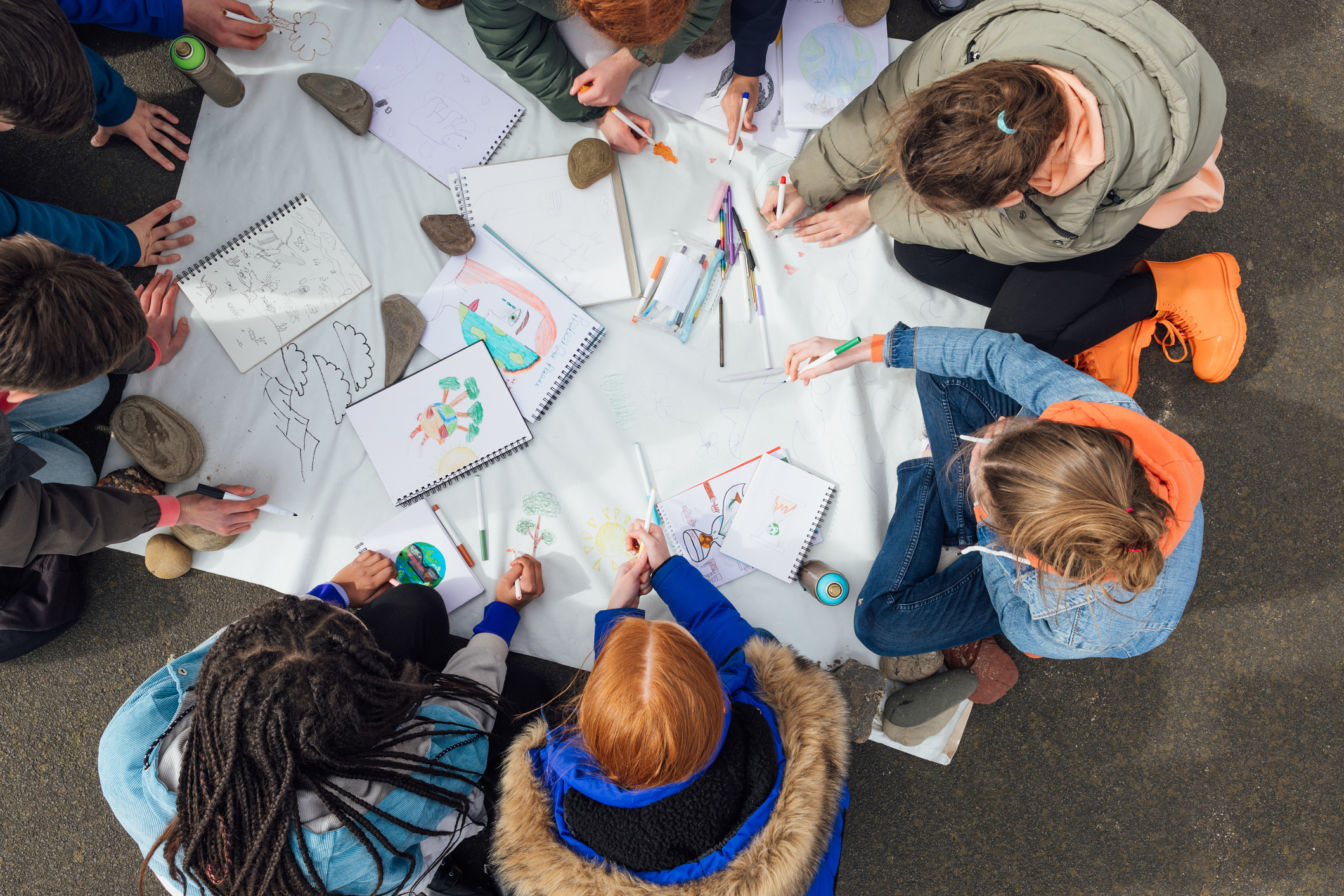 Children sitting outdoors on pavement drawing and coloring on large paper with markers and sketchbooks.