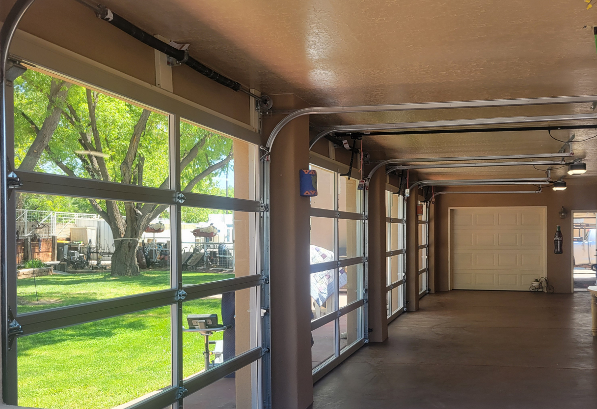 Interior view of a spacious garage with open glass-panel garage doors revealing a green lawn and tree outside, beige walls, ceiling lights, and a closed white garage door at the far end.