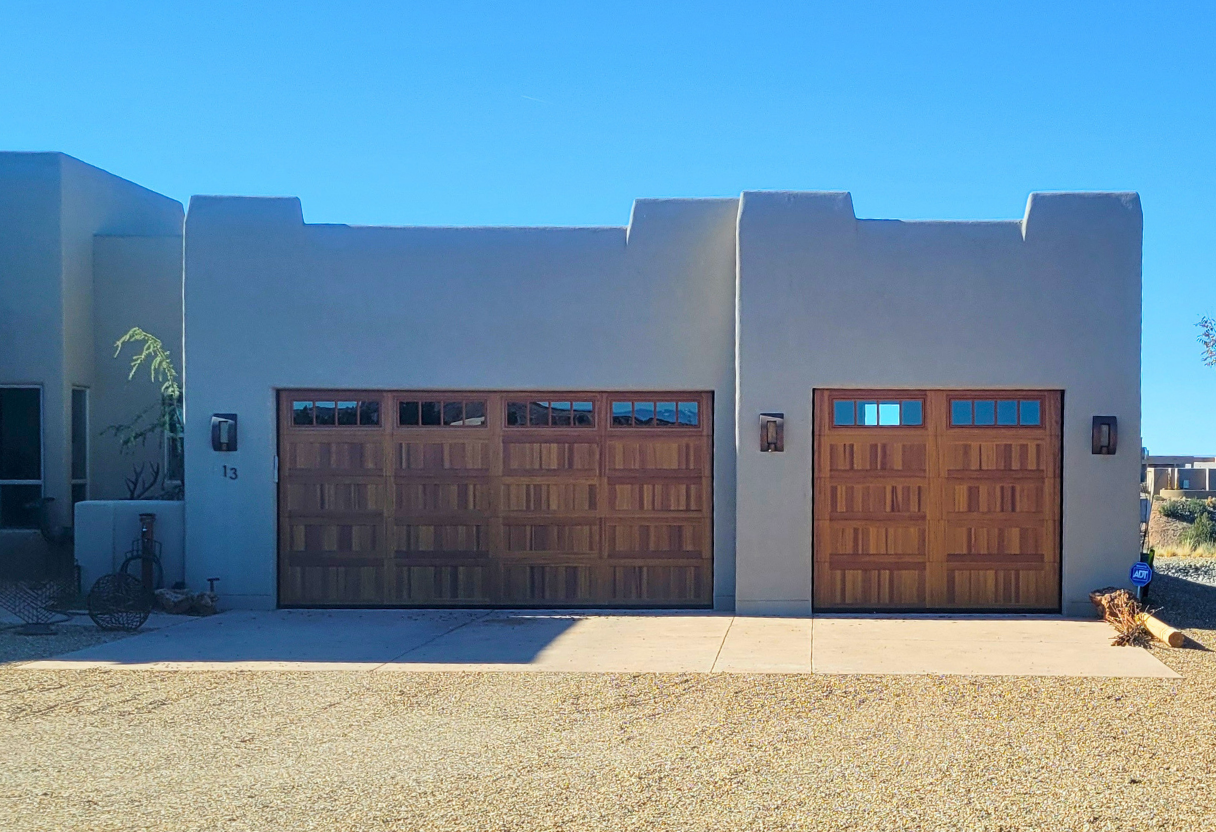 Two wooden garage doors on a single-story southwestern-style house with smooth beige stucco walls and a flat roof under a clear blue sky.
