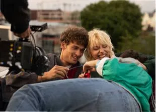 Two people smiling and looking at a phone while a third person leans forward in an outdoor setting.