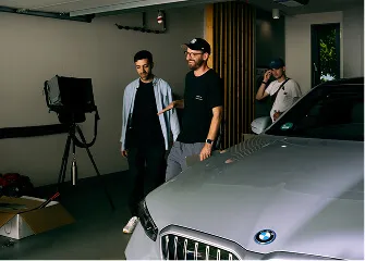 Three men in a garage setting near the front of a silver BMW car with studio lighting equipment in the background.