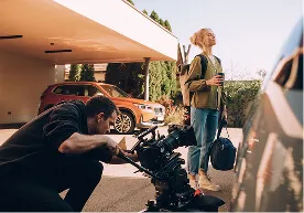 Videographer filming a woman standing outside near cars, holding a coffee cup and a suitcase.
