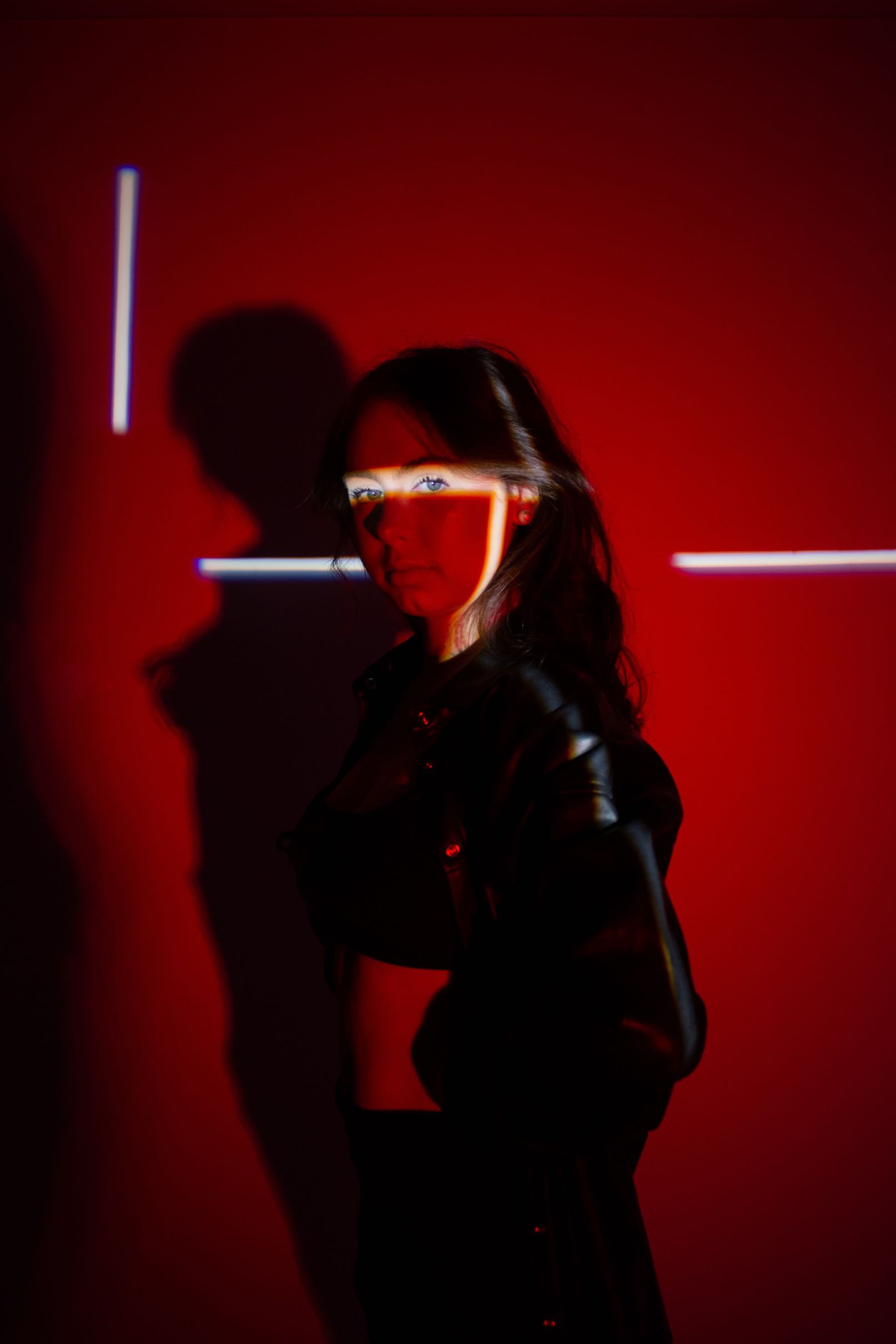 Woman with blue eyes standing against a red background with bright geometric light projections on her face and shadow behind.