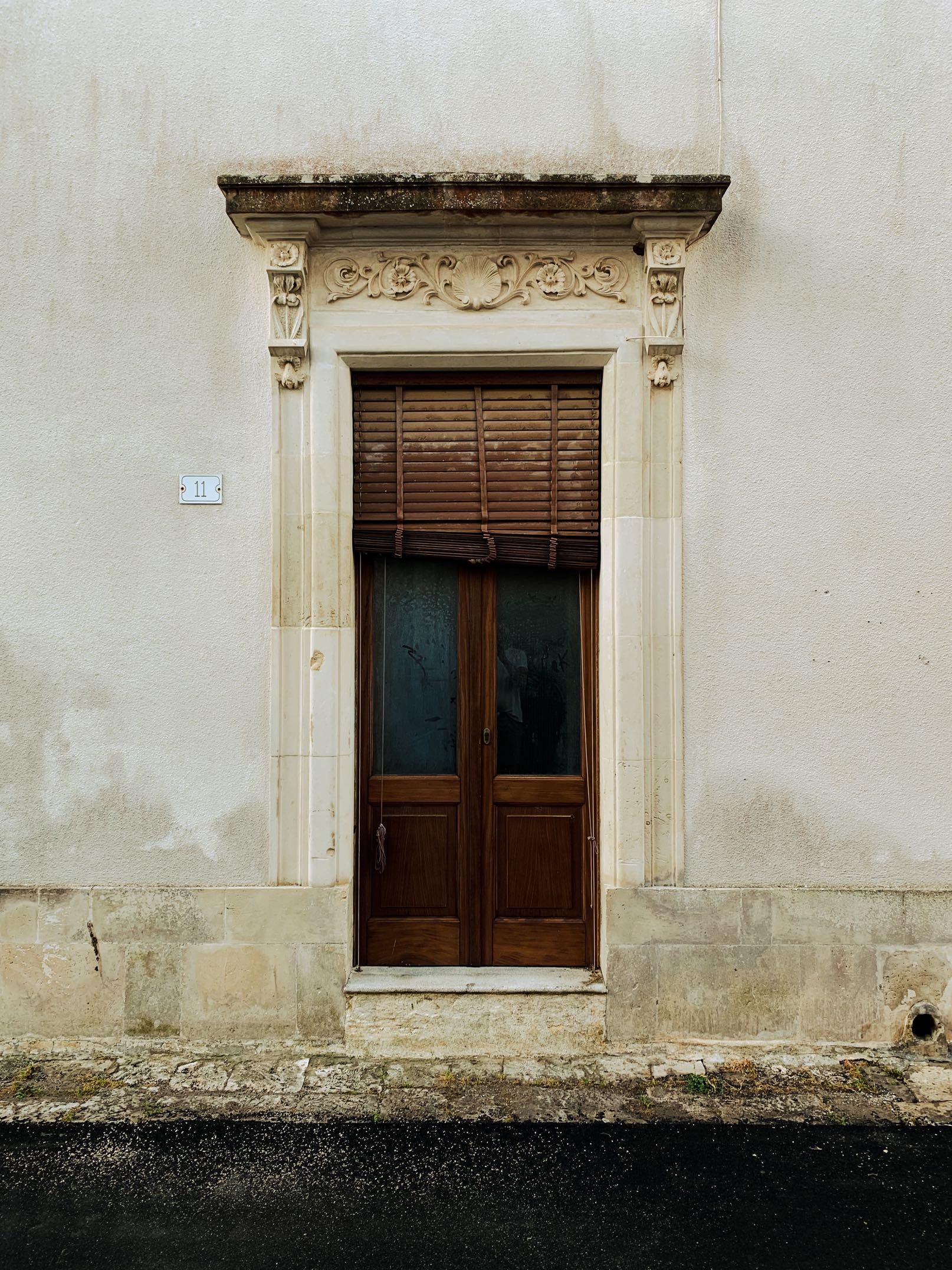Ornate wooden door with dark glass panels and partially closed brown blinds set in a beige stone frame on a weathered beige wall.