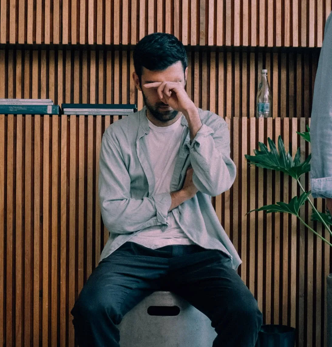 Man sitting on a stool against a wooden slat wall, rubbing his eyes with his hand.