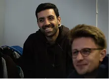 Two men indoors, one smiling directly at the camera wearing a black hoodie, the other wearing glasses and slightly out of focus.