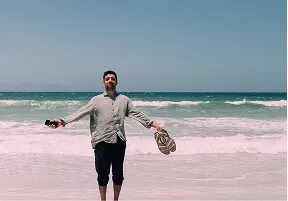 Man standing on the beach with arms outstretched holding sandals, facing the ocean under clear sky.