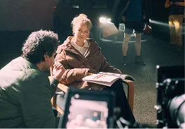 Woman sitting under bright light holding an open book, talking to a man indoors.