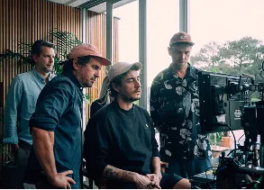 Four men intently watching a camera monitor indoors near a large window with trees visible outside.