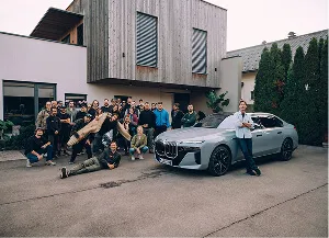 Group of people posing excitedly next to a silver luxury car parked outside a modern building.