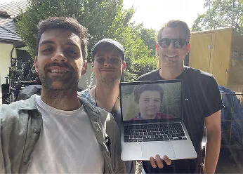 Three men outdoors smiling, with the man on the right holding a laptop showing a boy's photo on the screen.