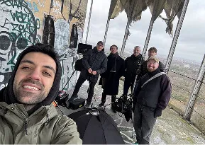 Group of six men standing on a rooftop with graffiti walls and a city view in the background.