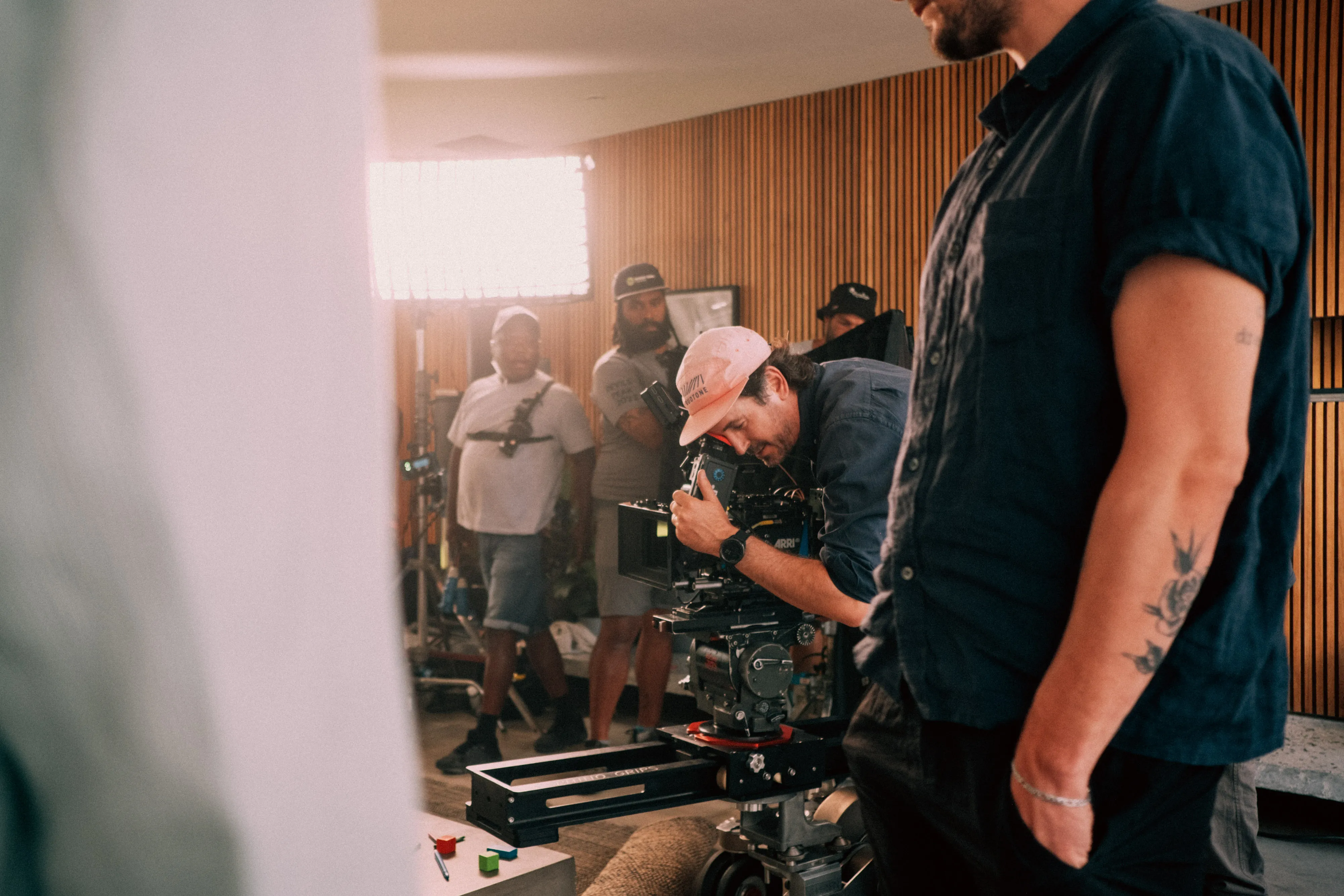 Film crew setting up camera and lighting inside a room with wood panel walls.