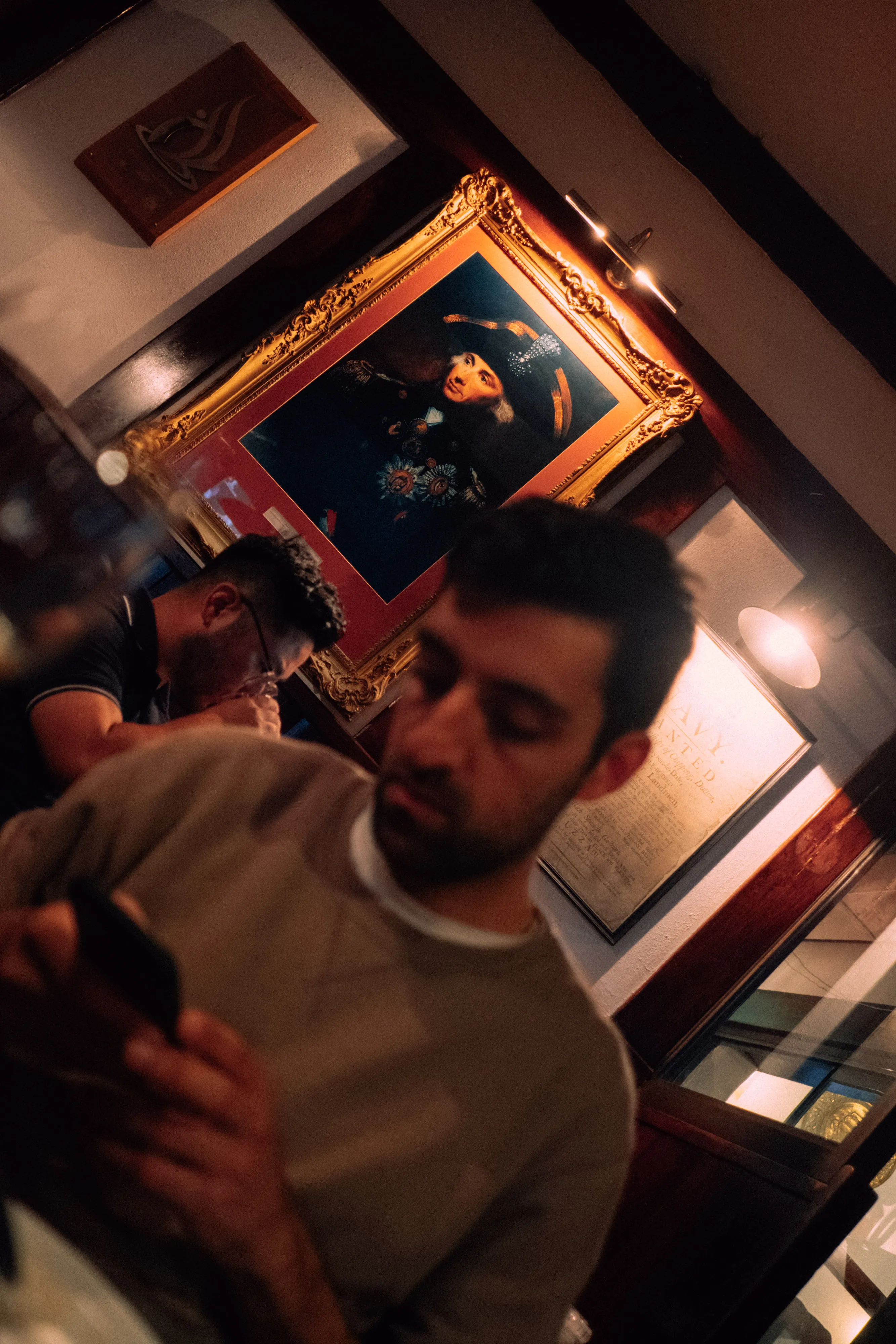 Two men seated indoors, one using a phone, with a framed historic portrait and document on the wall behind.