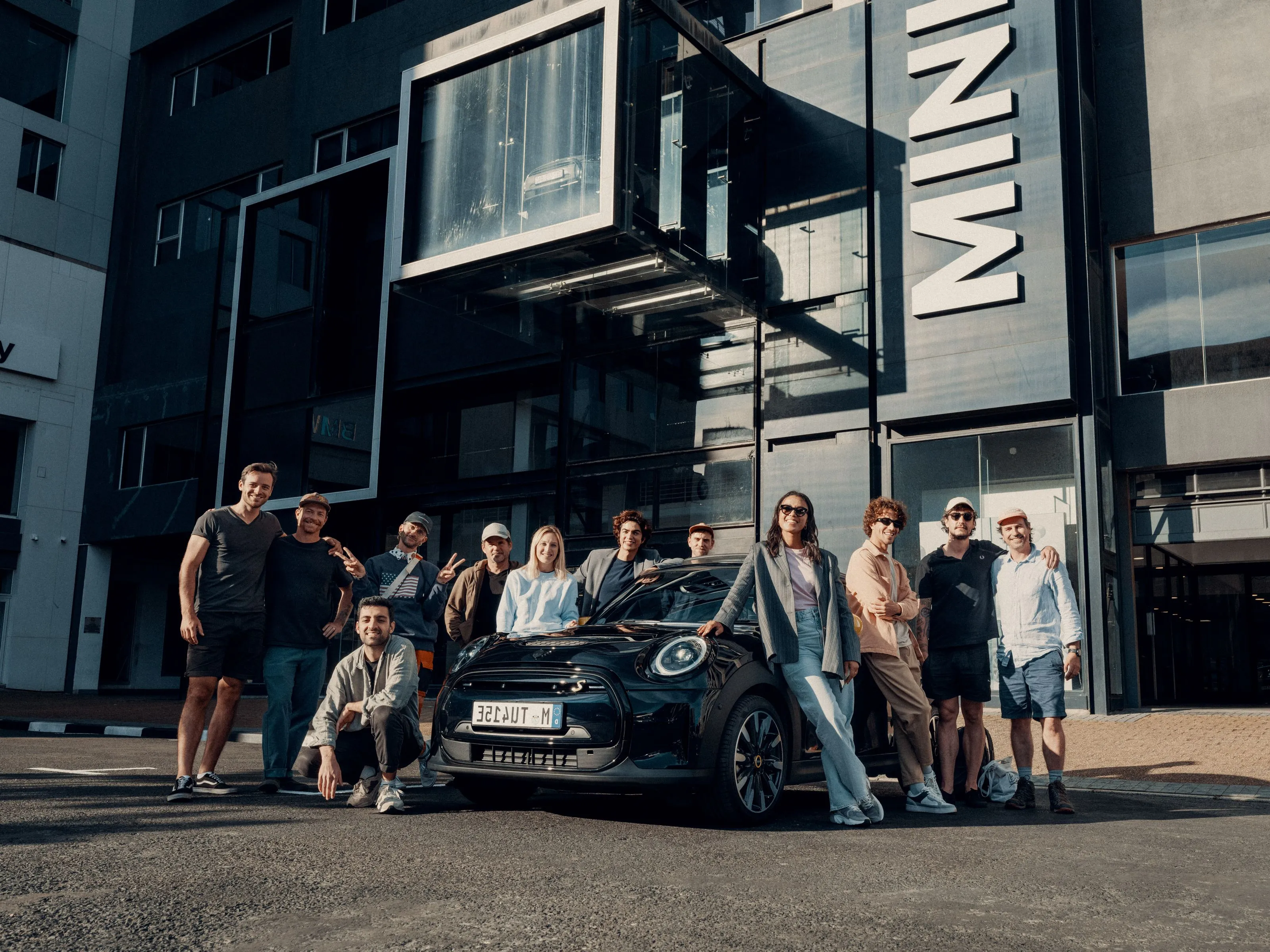 Group of people pose around a black Mini Cooper car outside a modern glass building with Mini signage.