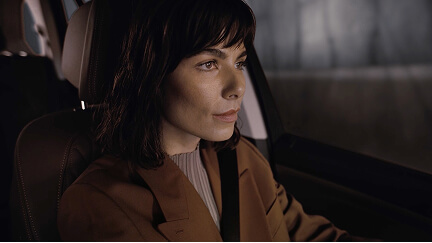 Woman with short dark hair wearing a seatbelt and brown coat driving a car.