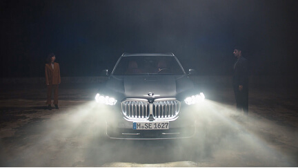 Front view of a BMW car with headlights on and two people standing on each side at night.