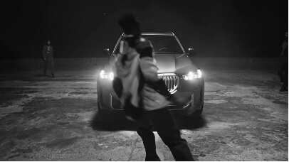 A person in motion in front of a car with headlights on at night, with two people in the background.