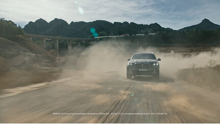 SUV driving on a dusty dirt road with mountains and a bridge in the background.