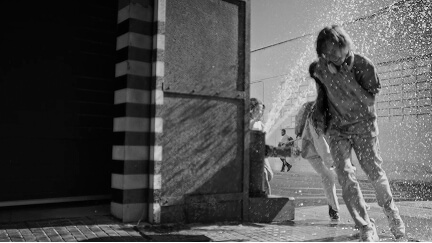 Two children playing and running through water spray on a street, shown in black and white.