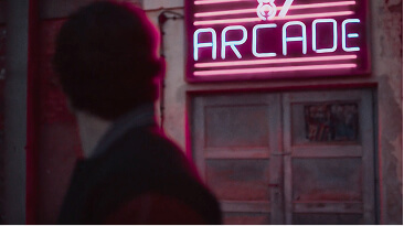 Silhouetted person looking at a glowing pink neon arcade sign on a brick wall at night.