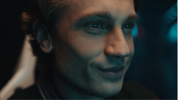 Close-up of a thoughtful young man with blue eyes in soft lighting and a dark background.