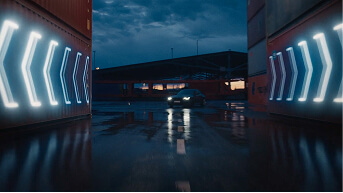 Car driving through a wet industrial area at night, flanked by glowing blue arrow lights on walls.