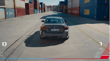 Rear view of a gray car driving through a shipping container yard on a sunny day.