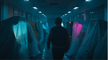 Person walks down a dim hallway with objects covered in white cloth and blue and pink lighting.