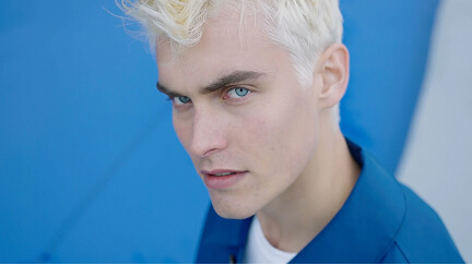 Portrait of a young man with platinum blonde hair and blue eyes, wearing a blue blazer.