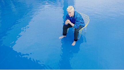Blond man in blue jacket sitting on a wire chair in shallow clear blue water, barefoot.