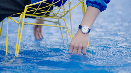 A person's hand with a watch touches clear blue water beneath a yellow metal chair leg.