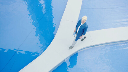Elderly woman with white hair walking on a white pathway surrounded by blue reflective water.