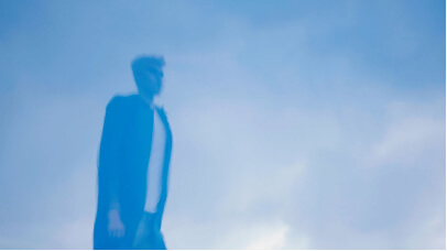 Blurred silhouette of a person walking against a blue sky with scattered clouds.