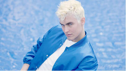 Young man with platinum blonde hair wearing a blue jacket over a white shirt against a blue background.