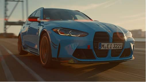 Blue BMW car driving on an open road at sunset with city skyline in the background.