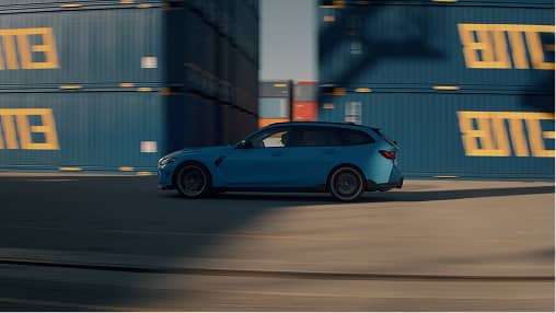 Blue station wagon driving past stacked blue shipping containers with yellow letters.