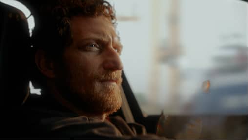 Bearded man with curly hair looking out a window with a thoughtful expression.